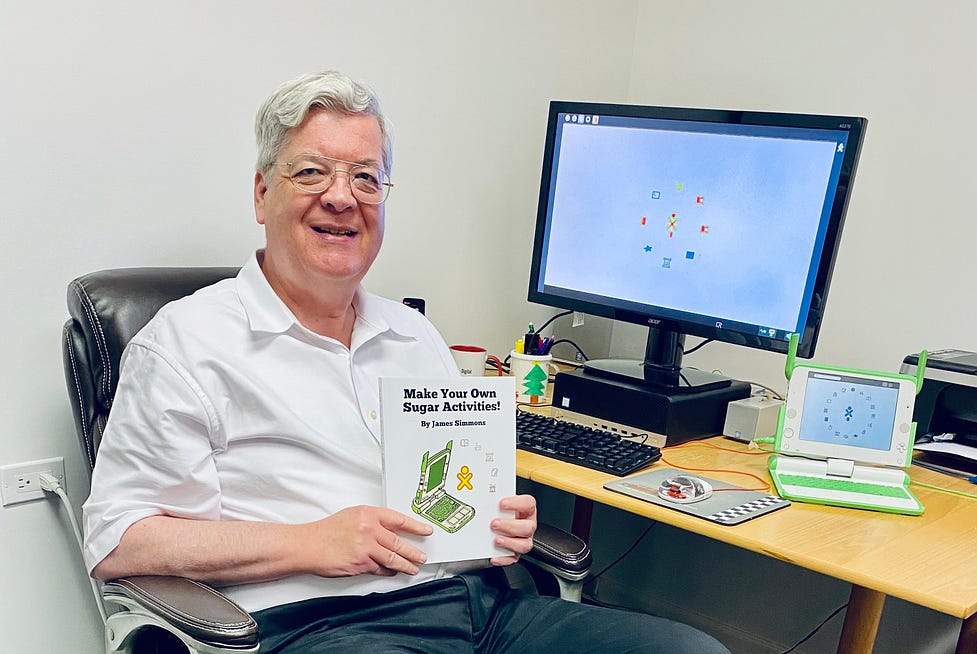 Photo of James Simmons holding a copy of “Make your own Sugar Activities”, sitting in front of a computer screen and a small OLPC laptop.