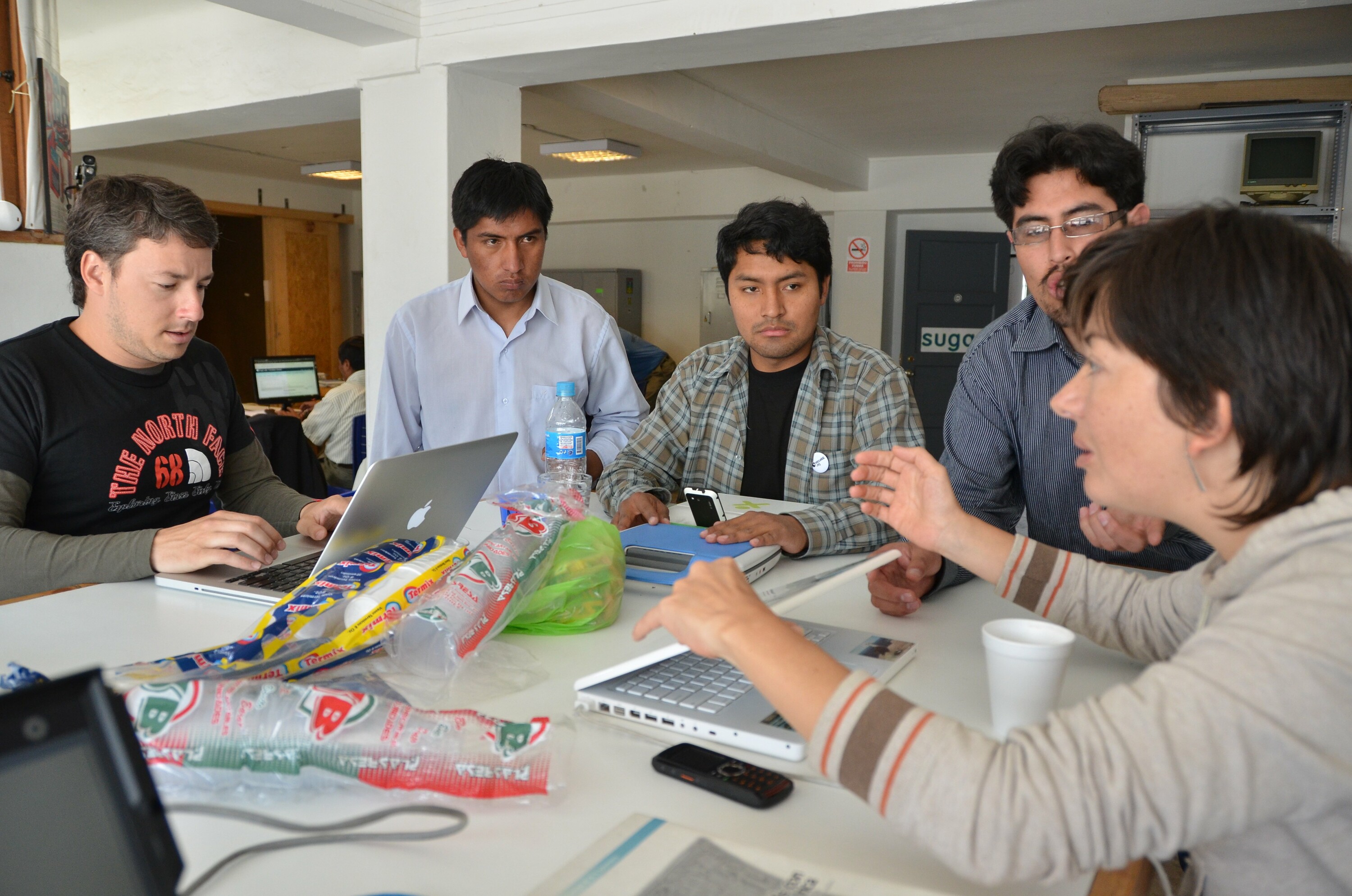 Five people sitting at a table together. You can see a variety of laptops on the table. The Sugar Labs logo is visible in the background, a sign attached to a door.